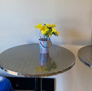 A small striped cup hold Shrpie maarkers with flowers attached to the ends to keep them in the store, sitting on a metal topped high round table.